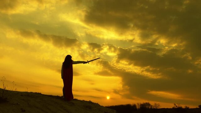 A super woman with a sword in her hand and in a red cloak stands on the mountain in rays of the sunset. Play Spartan. Free woman playing superhero. The girl plays a Roman in the sun.