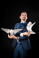 A magician with a smile in a blue tailcoat holds two white doves in his hands on a black background
