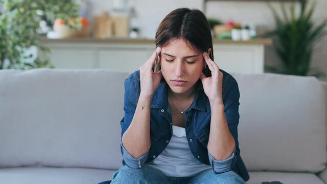Video of frustrated young woman with headache and frowning while sitting on couch in the living room at home.