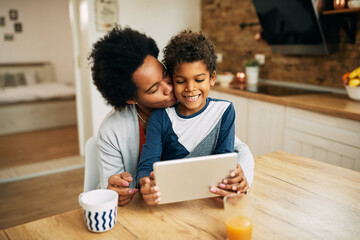 Affectionate black mother kissing her son while using using digital tablet at home.