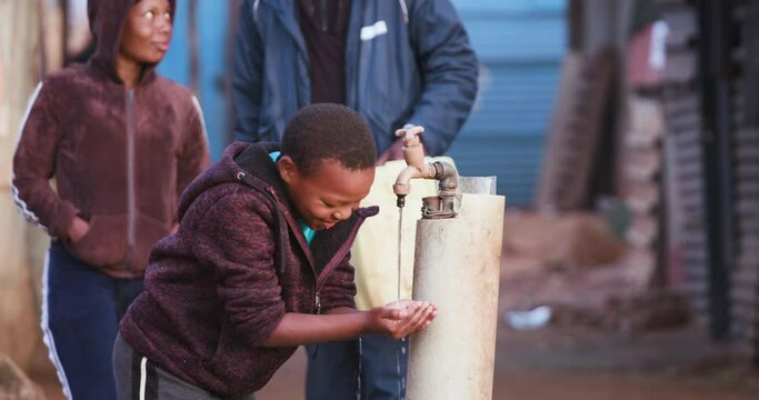 Water crisis. Close-up view of a young black african boy drinking water from a communal tap while people line up to collect water in plastic containers 