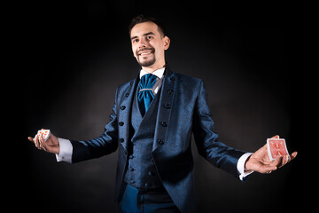 A magician in a blue dress coat on a black background holds cards in his hands