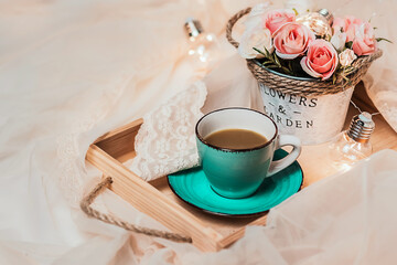 The atmosphere of a romantic morning, coffee in bed. Flowers in a vase and a cup of coffee on a wooden tray on a silk bed. Home interior. Lifestyle