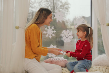 Happy mother and daughter decorating window with paper snowflakes indoors