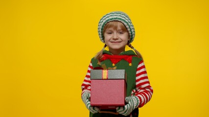 Little teen kid girl in Christmas elf Santa helper costume isolated on yellow background. Child giving present gift box to camera, smiling sweetly. People New Year holidays celebration