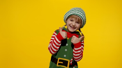 Teen joyful kid girl 5-6 years old in Christmas elf Santa helper costume smiling, showing thumbs up gesture isolated on yellow background. Child fooling around. People New Year holidays