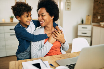 Happy African American boy embracing his mother who is working on laptop at home.