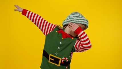 Little fun blonde kid teen teenager girl in Christmas elf Santa helper costume performing dab dance on yellow background. Child dancing making dabbing movement with hands up, internet meme gesture