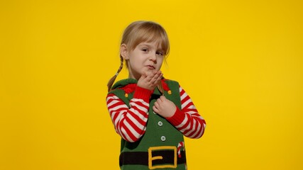 Naklejka premium Shy shamed child girl in Christmas elf Santa helper costume posing looking camera blows air kiss. Little kid feels guilty. Isolated on yellow background studio. People emotions. New Year holidays