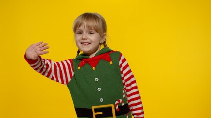 Fototapeta premium Teen kid girl 5-6 years old in Christmas elf Santa helper costume waving greeting with hand as notices someone. Hello, hi, greetings gesture. Small child laughing. People New Year holidays celebration
