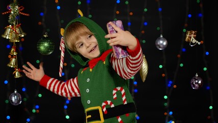 Kid girl in Christmas elf Santa Claus helper costume making selfie on smartphone near festive...