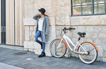 Fototapeta premium Portrait young pretty Caucasian girl, dressed in a gray jacket, a white sweater and a hat, leaning against the wall with a bike for a ride in the city, talking on the mobile phone