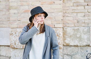 Portrait young pretty Caucasian girl, dressed in a gray jacket, a white sweater and a hat, talking on the mobile phone