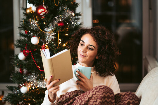 A brunette woman sitting on the sofa while reading a book in a cold day on December. She is near to a Christmas tree and she is covering herself with a blanket. Christmas holidays concept.