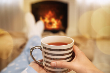 Woman with cup of tea resting near fireplace at home, closeup