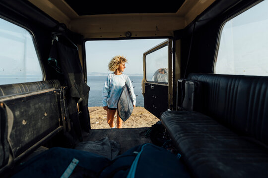 Woman holding surfboard while standing by car against sea at beach