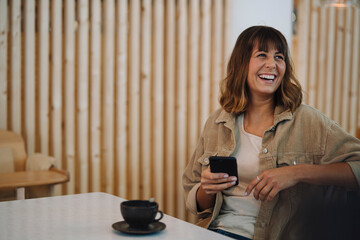 Female owner laughing looking away while using smart phone sitting in cafe