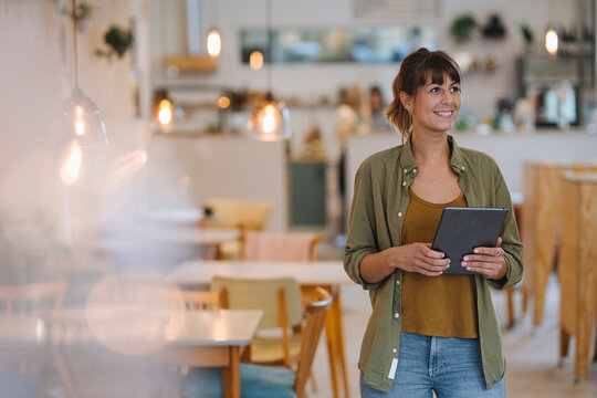 Smiling female entrepreneur looking away while holding digital tablet standing in cafe