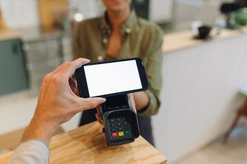 Cropped hand of customer doing contactless payment with smart phone while female owner holding credit card reader at checkout in coffee shop