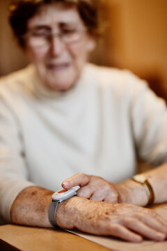 Wrinkled worried woman pressing emergency button on wrist over table at home