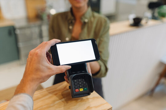 Cropped hand of customer doing contactless payment with smart phone while female owner holding credit card reader at checkout in coffee shop