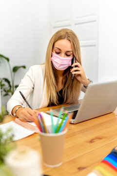 Blond woman wearing face mask writing in book while sitting at office