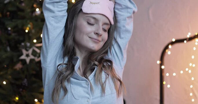 Xmas lazy morning, girl take off sleep mask, stretching on festive background of Christmas tree decorated with lights. High quality HD footage