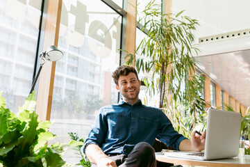 Cheerful young businessman looking away while sitting with laptop at table in cafe