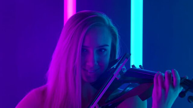 Smiling female musician masterly plays the violin in the studio. The blonde demonstrates her skill amidst the bright neon lights. Close up. Slow motion.