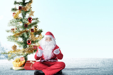 Asian man in Santa costume with open palm gesture sitting with Christmas tree