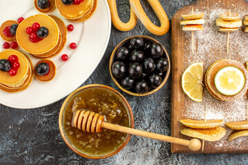 Fruit pancakes cookies near honey in a bowl and black cherries on gray table