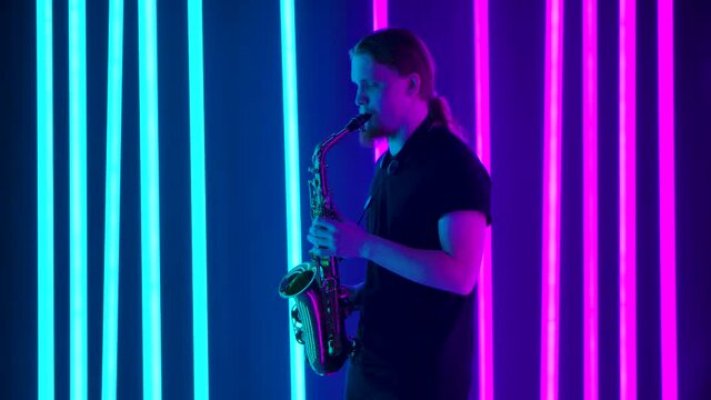 Side view of a young stylish man with long hair playing the saxophone masterly. The musician performs a live concert in a dark studio against the backdrop of bright neon lights. Slow motion.