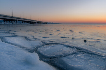 Ice-covered sea with cracks. The bridge is in the background. Dusk