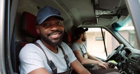 Close up of African American young happy handsome male courier sitting on front seat in van and smiling to camera while waiting for client with male colleague. Joyful delivery man in uniform