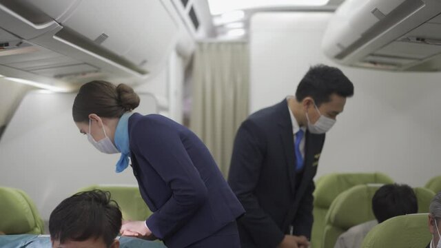 Caucasian female and Asian male flight attendants in a face mask for protective Covid-19 walk along aisle checking the passengers on safety standards with seat belt fastened before airplane take off.