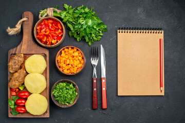 Tasty cutlets with vegetables with fork and knife next to notebook on dark background