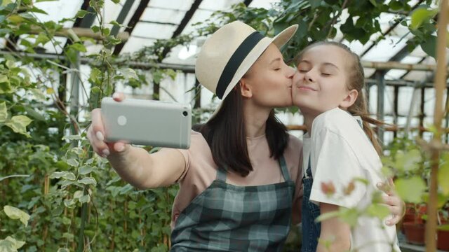 Slow motion of happy people mother and daughter taking selfie in greenhouse posing kissing smiling using smartphone camera. Gadgets and photography concept.