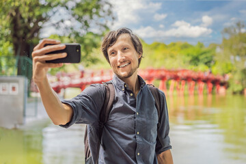 Fototapeta premium Caucasian man traveler on background of Red Bridge in public park garden with trees and reflection in the middle of Hoan Kiem Lake in Downtown Hanoi. Vietnam reopens after coronavirus quarantine COVID