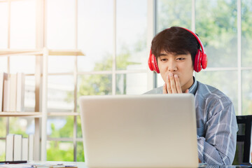 Asian young businessman happiness with red headphones he sitting on desk workplace home office looking to laptop computer monitor, confident handsome man lifestyle smile relax he listens to music