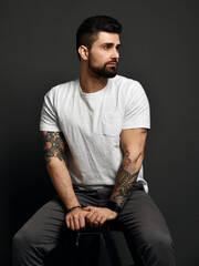 Handsome modern brutal bearded muscular man in t-shirt and jeans and with tattoo on arms sits with...