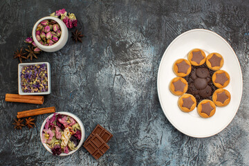 Fototapeta na wymiar top view of cookies on white plate and bowls of dry flowers and chocolates on grey ground