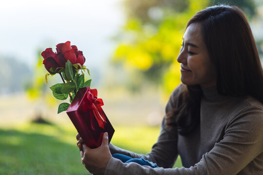 Closeup image of a beautiful asian woman holding red roses flower and a gift box in the park
