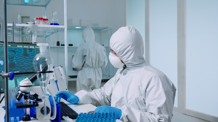 Scientist in ppe suit holding petri dish while studying virus samples in biotechnology lab typing on pc. Doctors examining vaccine evolution using high tech researching diagnosis against covid19 virus