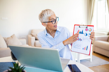 Focused businesswoman presenting charts and graphs on video call online. business, technology and people concept, Mature businesswoman with papers having video conference by laptop computer at office