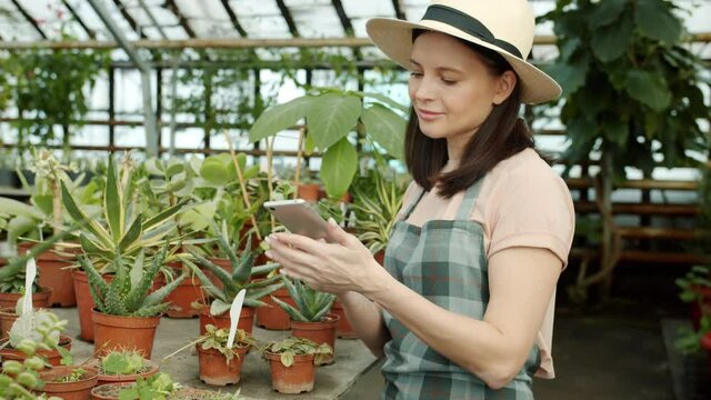 Young woman gardener and blogger is taking pictures of plants in greenhouse using smartphone camera and touching screen. People and gardening concept.