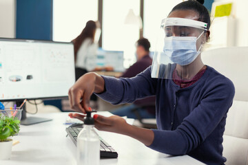African woman following safety precaution during covid19 using hand antibacterial. Businesswoman in new normal workplace disinfecting while colleagues working in background.
