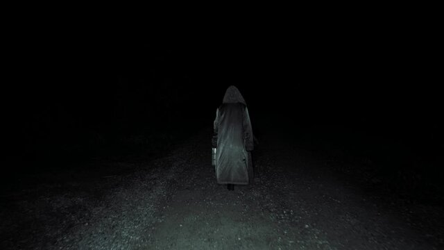 Old witch walking down dark spooky road. Scary old woman moving through darkness down gravel path in pitch black night time.
