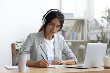 Distant learning. Confident woman student studying online making notes of lecture sitting by pc screen in earphone set. Focused young lady remote worker writing up client order received by video call