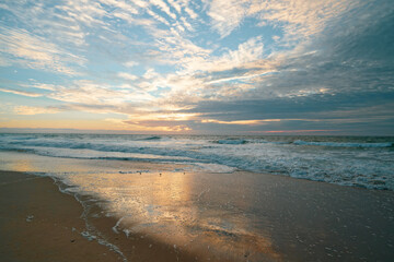 Sunset on the beach in light blue, yellow and pink colors. Empty sand beach, ocean waves, cloudy sky, and beautiful sun reflection on the beach