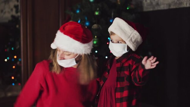 coronavirus, covid-19 in the new year. little daughter with her mother in a bad Santa hat and medical mask waving their hands at home under the Christmas tree.